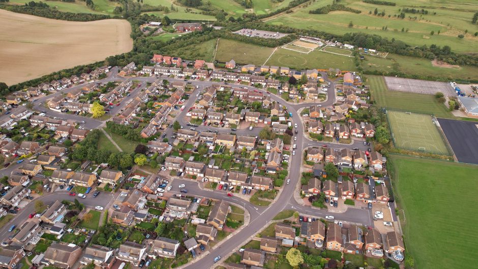 A detailed aerial view of a residential neighbourhood showing a variety of single-family houses, mostly with brown or grey tiled roofs and small gardens, arranged along a network of curved and straight streets. Numerous cars are parked in driveways and along the streets, with some parked parallel and others in designated parking areas. The surrounding environment includes green spaces, including several sports fields with green playing surfaces and visible boundary markings, situated to the right of the residential area. In the background, open fields, patches of woodland, and occasional farm buildings extend towards the horizon, indicating a semi-rural setting. The lighting suggests daytime with overcast or partly cloudy conditions, highlighting the textures of rooftops and foliage. This scene exemplifies a typical suburban area, which could benefit from independent waste collection services offered by Rubbish Removal Sutton, aligning with the context of private waste handling and rubbish removal services for local homes in the Cheam SM3 area.