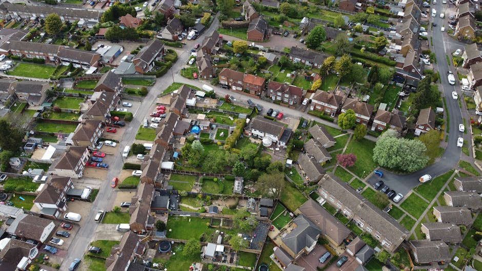 A detailed aerial view of a residential neighbourhood showing a variety of single-family houses, mostly with brown or grey tiled roofs and small gardens, arranged along a network of curved and straight streets. Numerous cars are parked in driveways and along the streets, with some parked parallel and others in designated parking areas. The surrounding environment includes green spaces, including several sports fields with green playing surfaces and visible boundary markings, situated to the right of the residential area. In the background, open fields, patches of woodland, and occasional farm buildings extend towards the horizon, indicating a semi-rural setting. The lighting suggests daytime with overcast or partly cloudy conditions, highlighting the textures of rooftops and foliage. This scene exemplifies a typical suburban area, which could benefit from independent waste collection services offered by Rubbish Removal Sutton, aligning with the context of private waste handling and rubbish removal services for local homes in the Cheam SM3 area.
