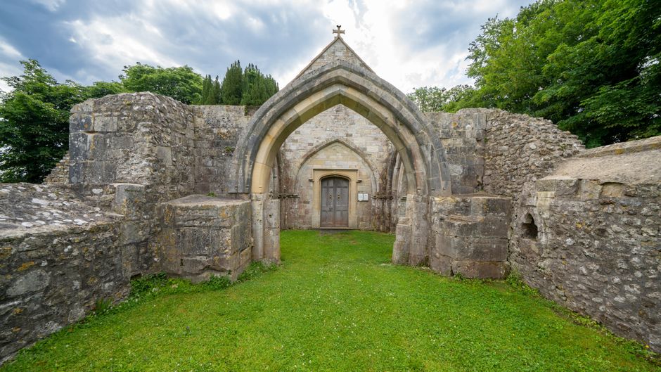 An ancient stone structure with a pointed Gothic arch entrance, constructed from weathered light gray stones, stands amidst a grassy area. The archway features detailed moldings and is framed by thick, uneven stone walls that extend outward on both sides, with some portions appearing partially crumbled or eroded. The wooden door within the entrance is dark brown with decorative metal hinges and a small rectangular plaque beside it. Surrounding the structure, lush green trees and foliage are visible, indicating a park or historic site setting. The sky overhead is partly cloudy, with patches of blue peeking through. The scene is illuminated by natural daylight, emphasizing the texture of the stonework and the vibrant greenery. The overall environment suggests a preserved historic building, possibly a former church or monastic gateway, situated in an outdoor landscape that may be associated with heritage or archaeological sites, aligning with themes of independent or private heritage site management related to waste removal or preservation services in the area.