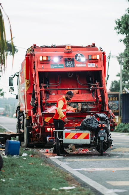 A waste collection worker wearing a high-visibility orange and yellow vest is standing on a paved area next to a large red rubbish truck, which is parked on the roadside. The back of the truck is open, revealing mechanical lifting components and a partially visible compartment for waste. The worker appears to be handling or inspecting black plastic garbage bags placed on a wheeled cart that is positioned behind the truck. The scene is set outdoors, with a grassy verge on the left side, a small section of curb, and a road leading into the distance. In the background, there are some trees, utility poles, and a faint indication of other vehicles or street signs, suggesting a residential or commercial area. The lighting is natural, indicating daylight conditions. This scene illustrates an independent or private waste collection activity, perhaps handled by a local rubbish removal service such as Rubbish Removal Sutton, with an emphasis on on-site rubbish disposal and waste management services.