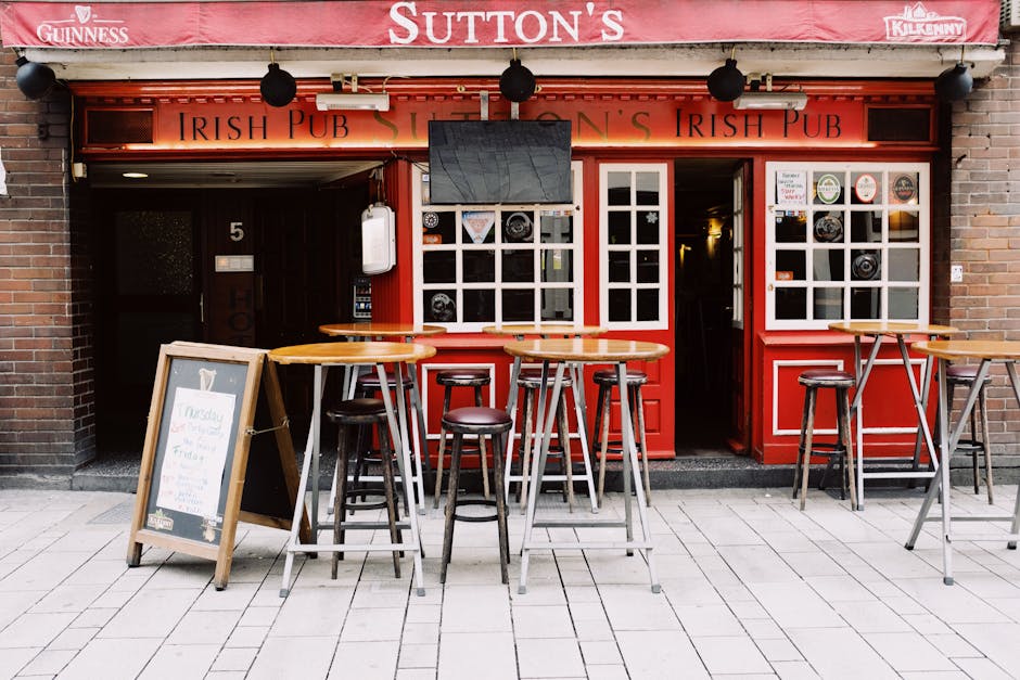 The image depicts the exterior of a traditional Irish pub located on Sutton High Street, with a red-painted facade and branding that includes signage for Guinness and Kilkenny. The pub has a large windowpane with white framing, revealing a warmly lit interior, and a central open doorway inviting patrons inside. In front of the pub, there are several high wooden tables with rounded tabletops and dark brown bar stools, arranged on a light grey tiled pavement. The tables and stools are positioned to facilitate outdoor seating, and one small chalkboard sign stands on an easel at the left side, partially obscured, likely displaying specials or menu information. The pub's frontage features hanging black outdoor lights, and a large flat-screen television is mounted above the window, angled downward for visibility from outside. The surrounding environment suggests a lively street scene with the pub ready to welcome customers, and the overall setting aligns with a typical UK social venue amidst a busy high street, where alternative waste handling or rubbish collection services by Rubbish Removal Sutton could be relevant for maintaining cleanliness and hygiene in such commercial premises.
