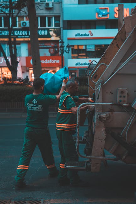 A waste collection worker wearing a high-visibility orange and yellow vest is standing on a paved area next to a large red rubbish truck, which is parked on the roadside. The back of the truck is open, revealing mechanical lifting components and a partially visible compartment for waste. The worker appears to be handling or inspecting black plastic garbage bags placed on a wheeled cart that is positioned behind the truck. The scene is set outdoors, with a grassy verge on the left side, a small section of curb, and a road leading into the distance. In the background, there are some trees, utility poles, and a faint indication of other vehicles or street signs, suggesting a residential or commercial area. The lighting is natural, indicating daylight conditions. This scene illustrates an independent or private waste collection activity, perhaps handled by a local rubbish removal service such as Rubbish Removal Sutton, with an emphasis on on-site rubbish disposal and waste management services.